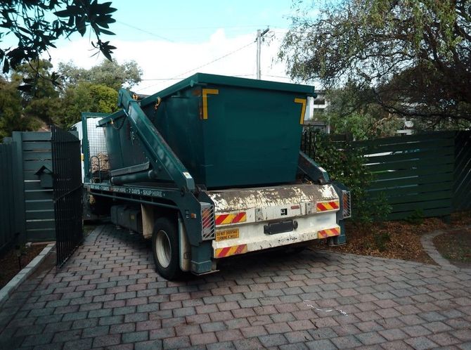 Green garbage truck parked in a driveway beside dark green fencing and trees
