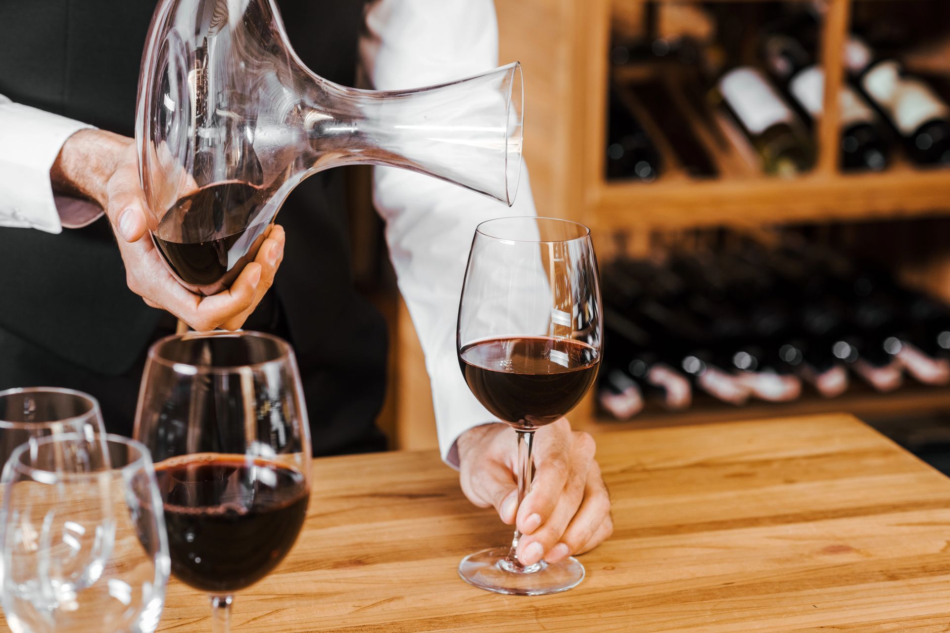 Un sommelier travasa il vino rosso in un bicchiere, altri bicchieri pieni sono sul bancone di legno. Sullo sfondo, un portabottiglie.