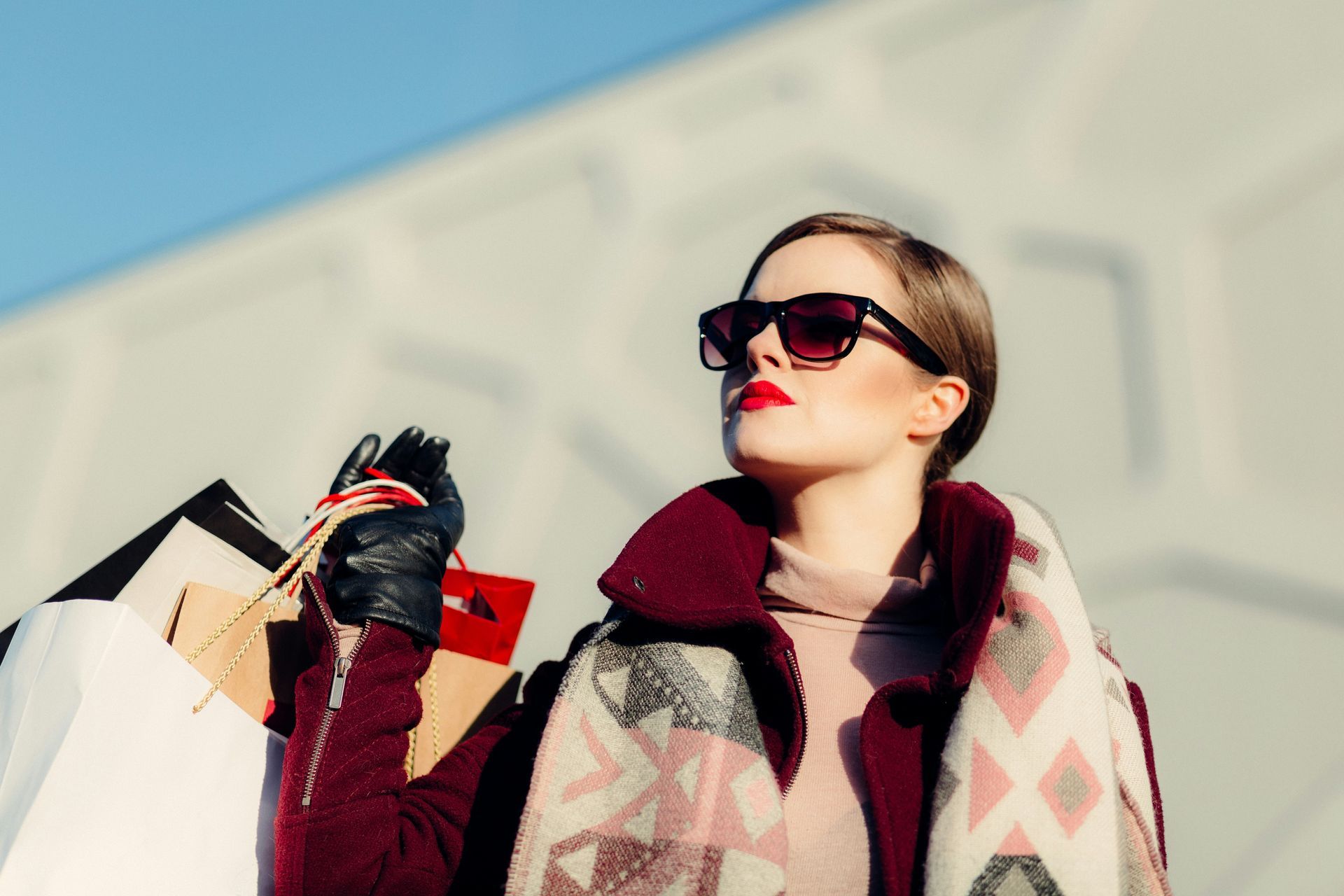 A person wearing sunglasses and a burgundy coat carries shopping bags in front of a geometric wall.