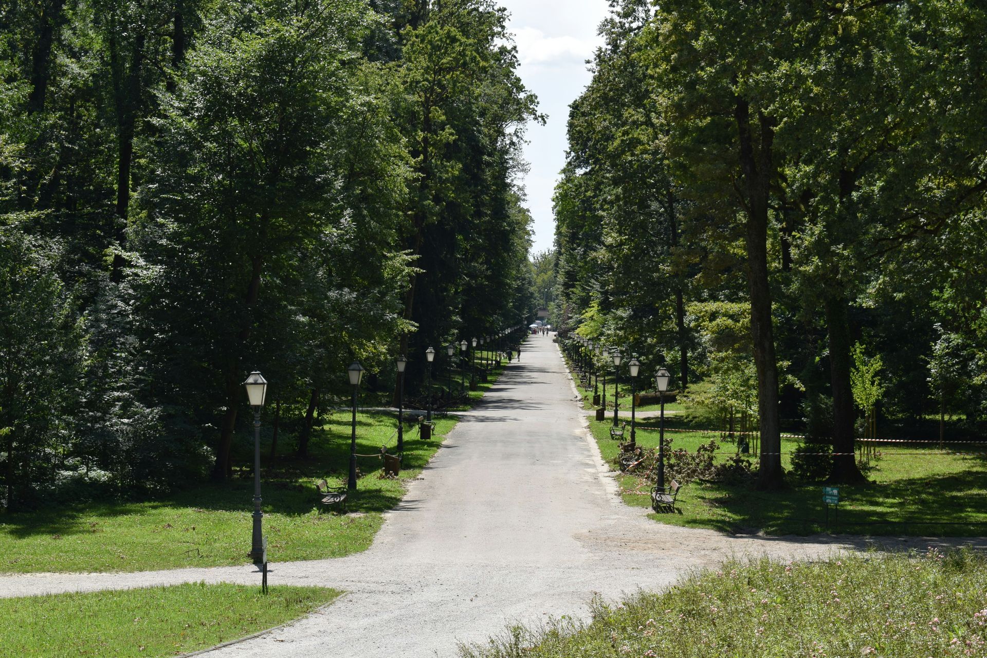 A gravel path lined with trees and lamp posts stretches into the distance through a green, sunlit park.
