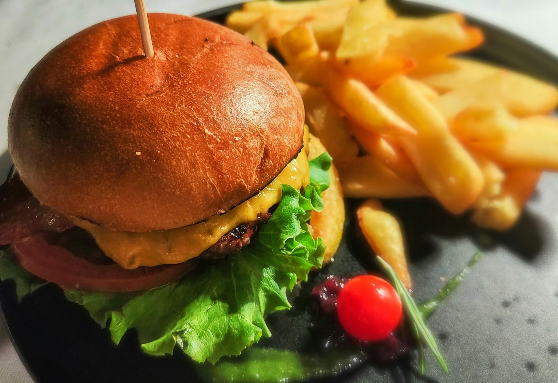 A cheeseburger with lettuce and a side of golden French fries on a black plate, garnished with a single cherry tomato.