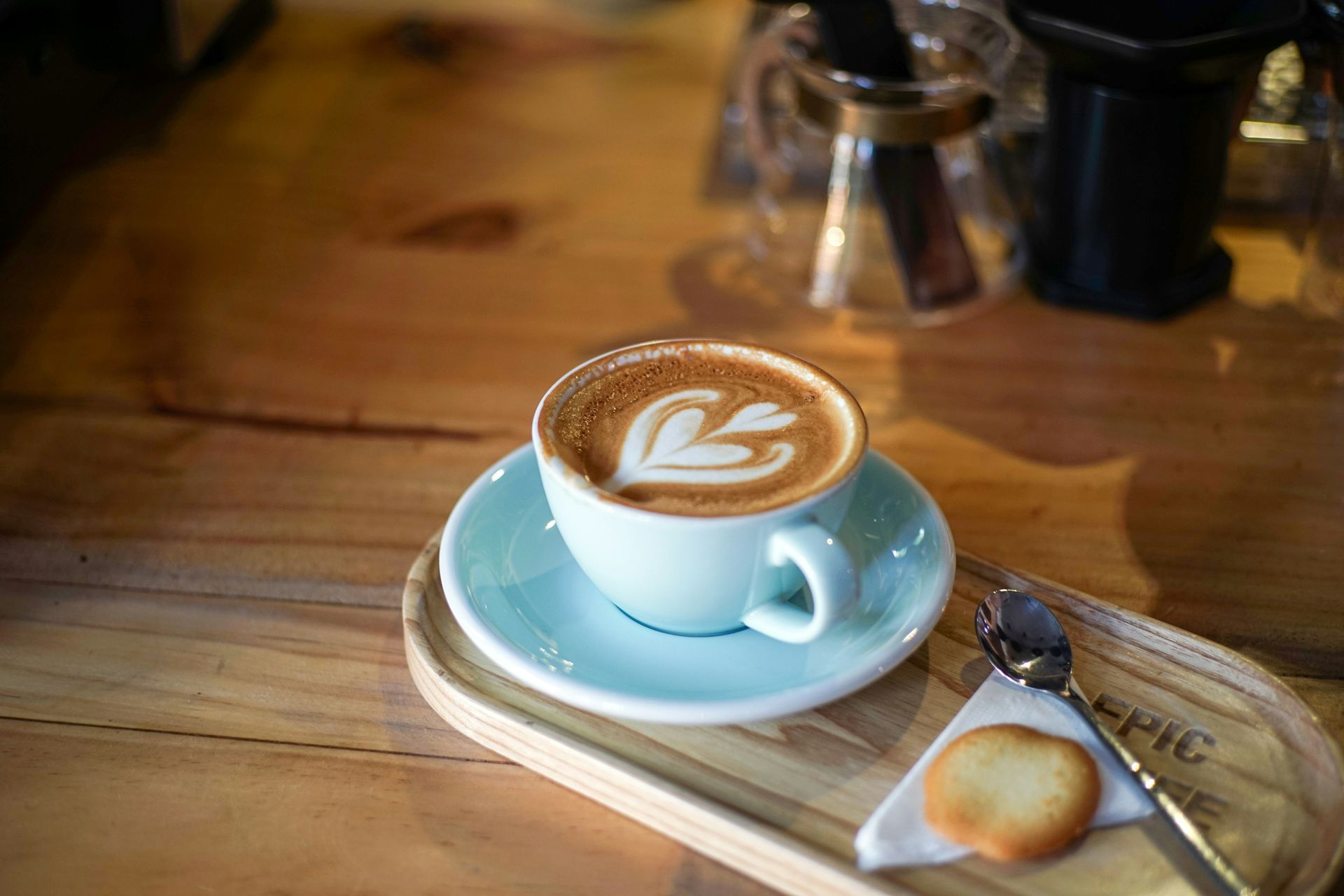A light blue cup of latte art coffee with a side cookie and spoon on a wooden tray, set on a rustic wooden table.