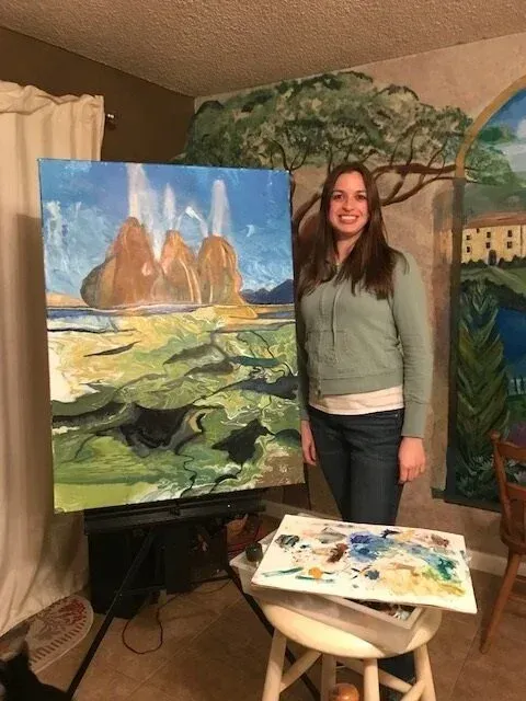 Woman standing beside a painting of rocky mountains, with a paint palette and easel in a studio.