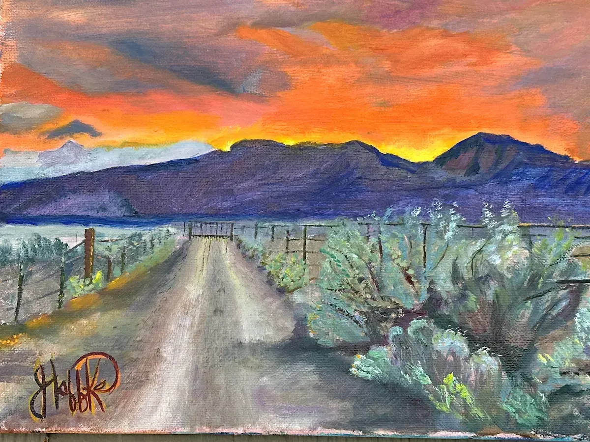 Desert road leading toward purple mountains under a vivid orange sunset sky, with shrubs and fence posts.
