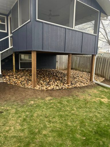 A dark blue screened-in porch elevated on wooden posts above a patch of ground covered in decorative stones and mulch.
