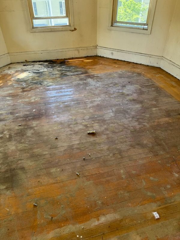 Empty room with worn wooden floor, patchy stains, and two windows letting in daylight.