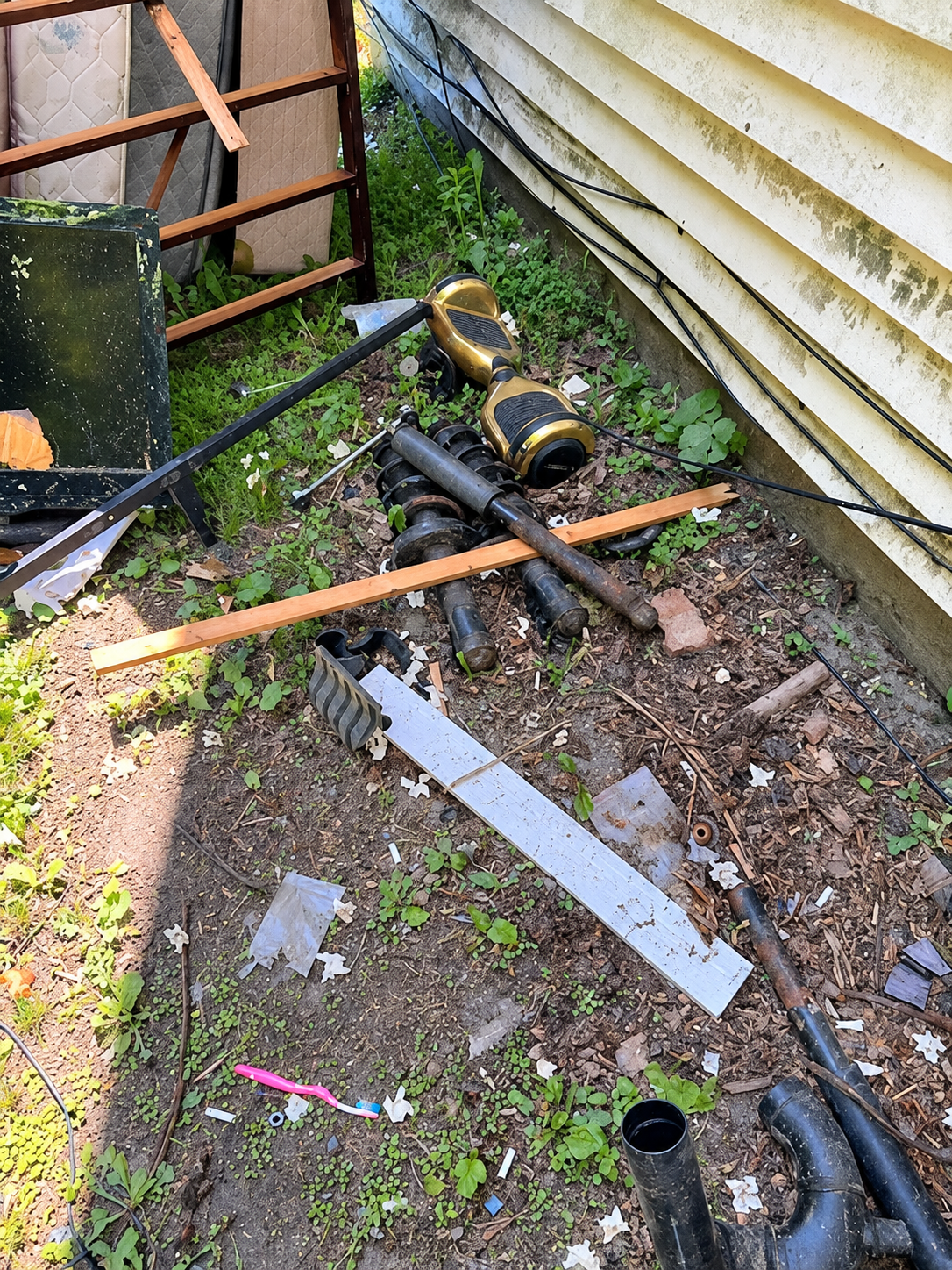 Debris-strewn narrow side yard with pipes, wood planks, and metal scraps beside a house and fence.