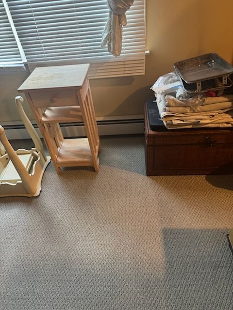 Small wooden side table by a window, next to stacked items on a chest and a white chair in a carpeted room