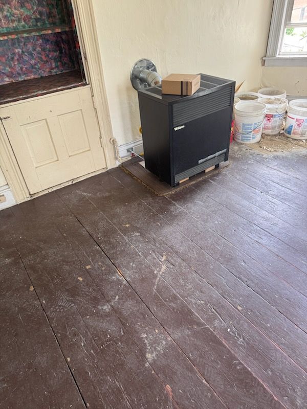 Empty room with worn hardwood floor, black cabinet by the door, and paint buckets by the window.