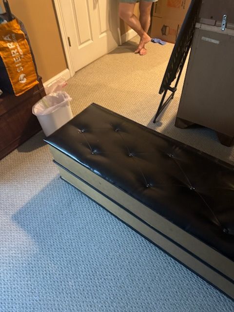 Black tufted storage bench in a carpeted room, with a trash bin and a person’s legs in the background.