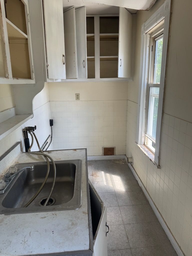 Empty kitchen with sink, white cabinets, tiled walls, and a window beside a narrow counter.