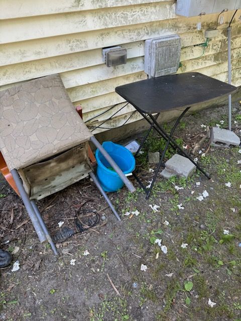 Old house siding with a tipped chair, folding table, blue bucket, and scattered debris on the ground