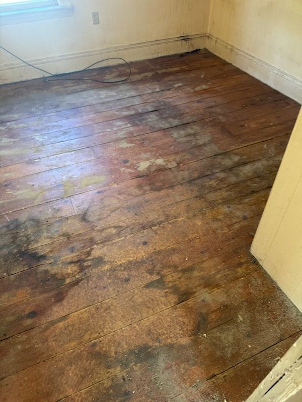 Bare room with worn, stained wooden floor and white walls, showing water damage.