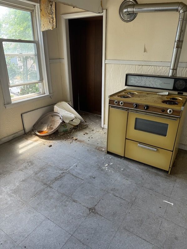 Abandoned kitchen with yellow stove, broken chair, peeling walls, and debris on the floor