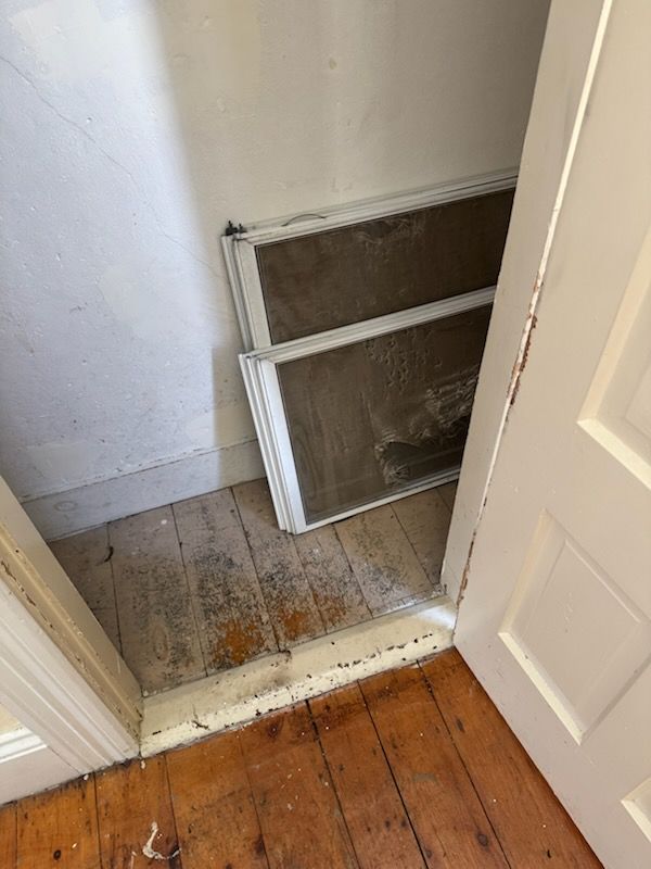 Two removed window screens leaning in a narrow doorway, with worn wooden floorboards and white walls.