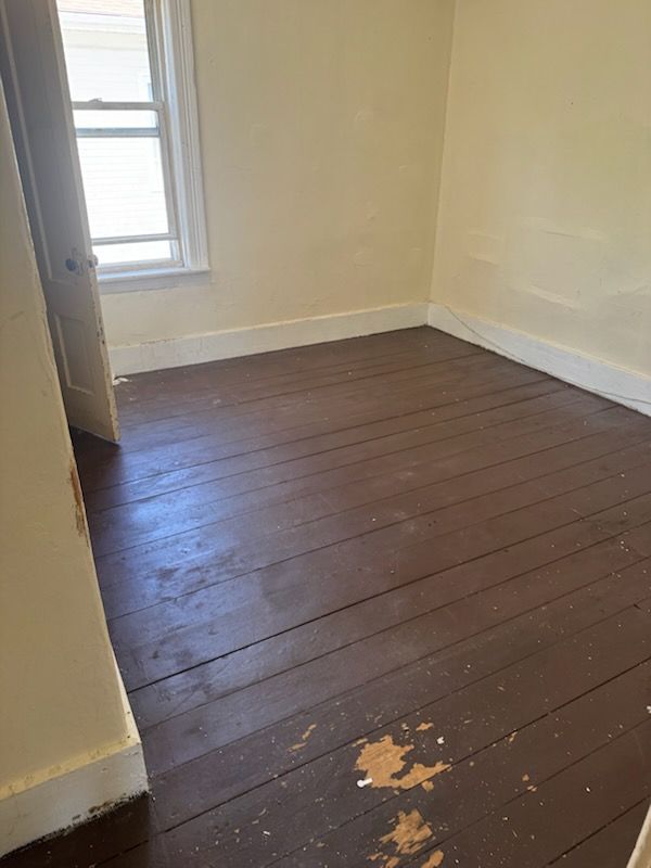 Empty room with dark wooden floor, chipped paint, and a window on the left.