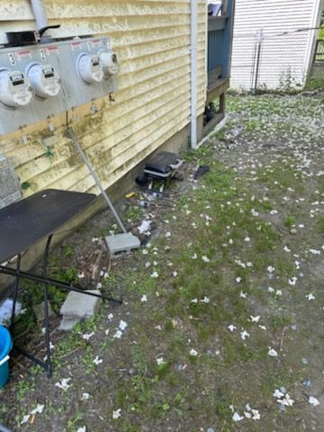 Side yard with gas meters on a yellow house, a black bench, and scattered debris on the ground