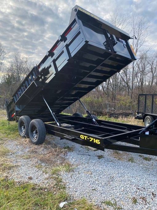 Black dump trailer raised steeply on a gravel lot under a cloudy sky