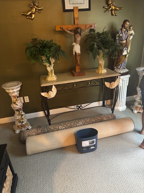 Home altar with crucifix, statues, plants, and a rolled carpet on the floor