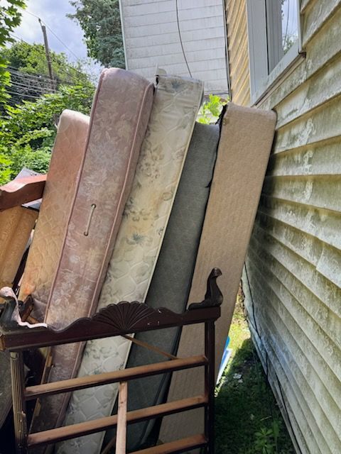 Mattresses and a wooden chair piled beside a house wall in a narrow outdoor passage.