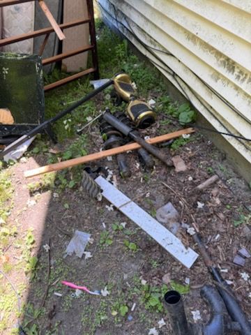 Cluttered yard corner with metal pipes, wooden planks, and debris beside a house wall and ladder