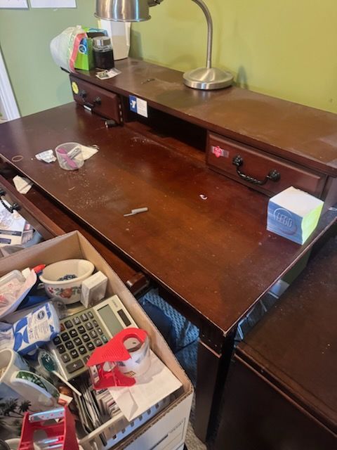 Cluttered wooden desk with drawers, lamp, tissue box, and office supplies in baskets