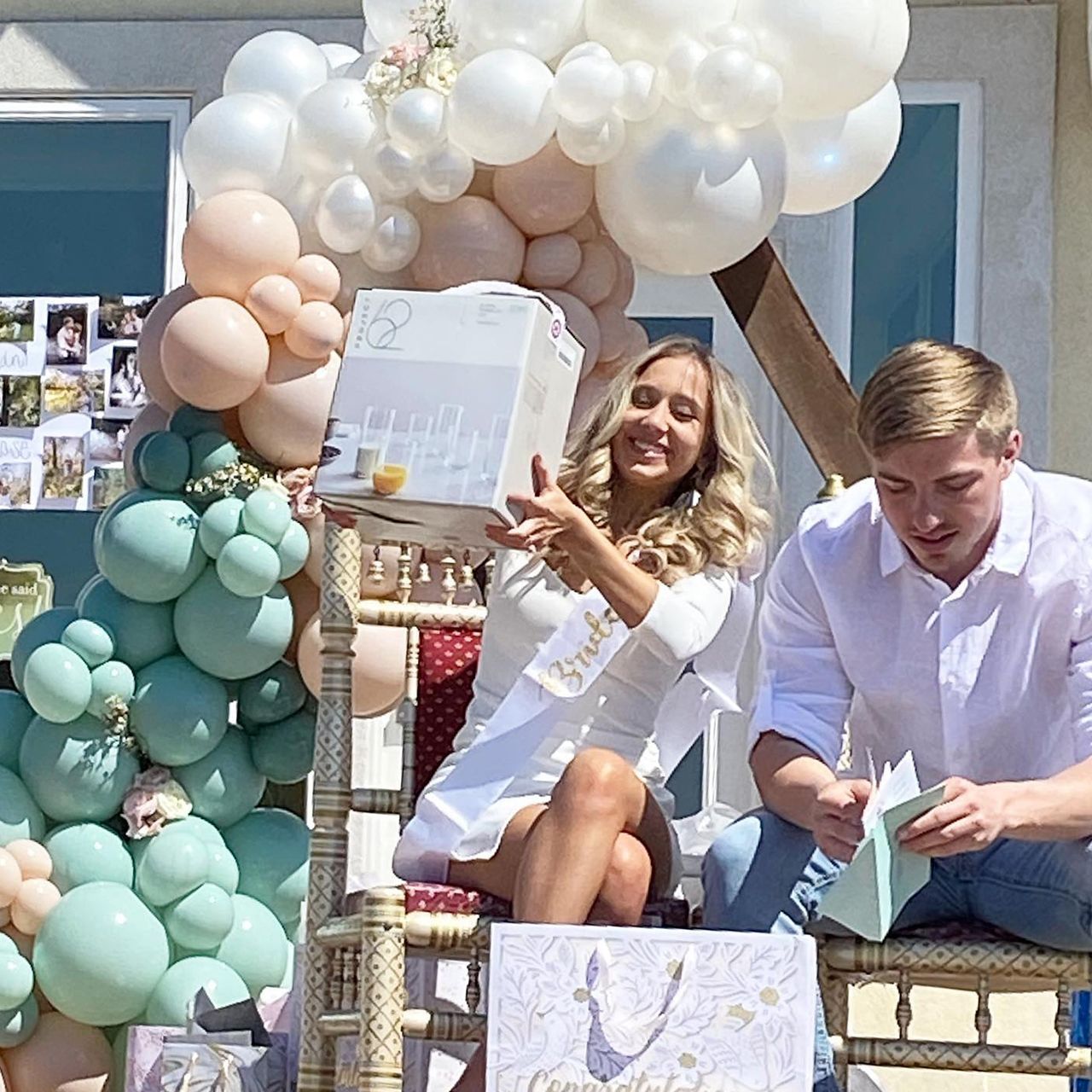 Woman in white dress smiles holding a gift box. A man sits beside her, opening a card, surrounded by balloons.