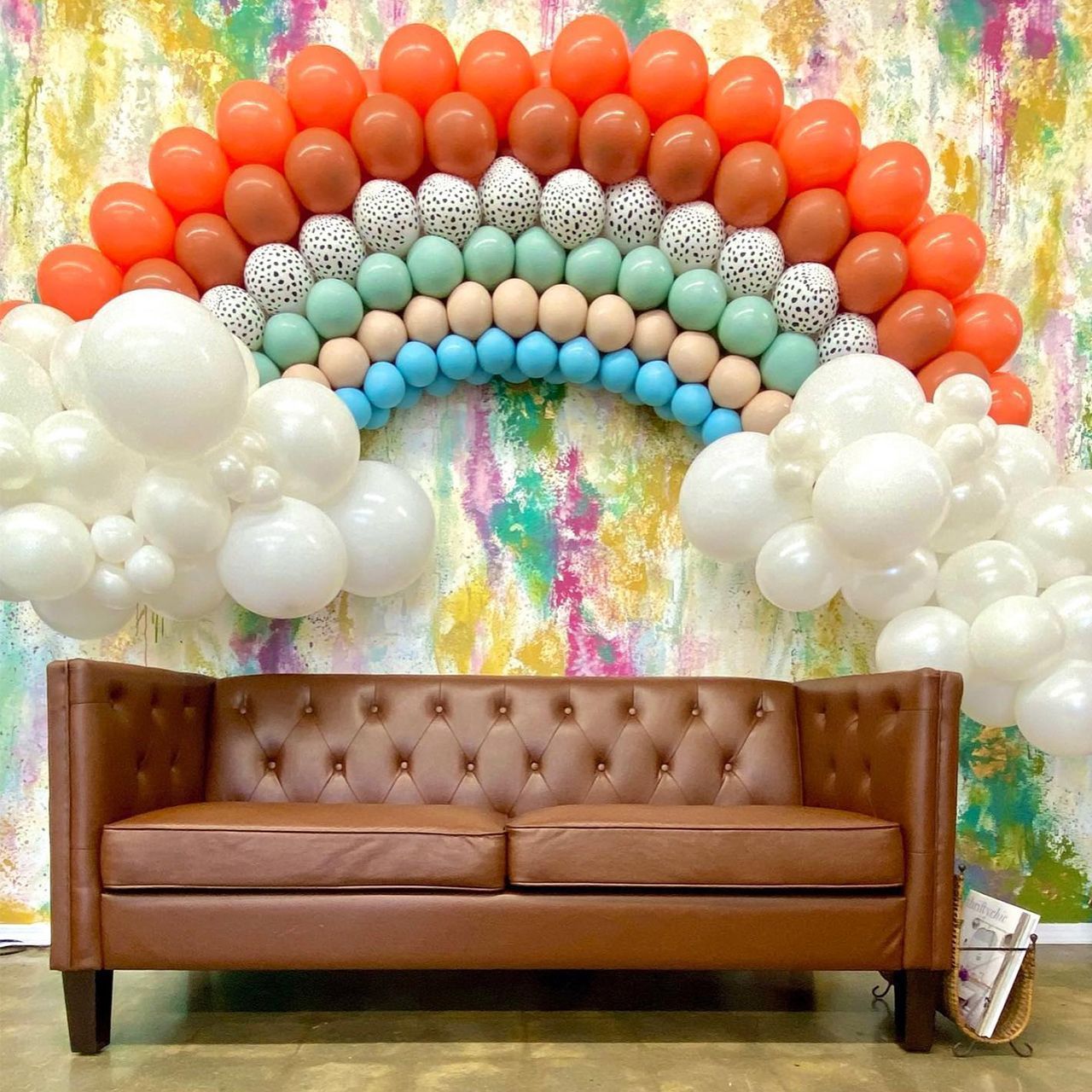 Brown sofa below a rainbow balloon arch against a colorful wall; white balloon clouds on the sides.