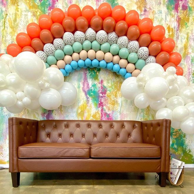 Brown sofa below a rainbow balloon arch against a colorful wall; white balloon clouds on the sides.
