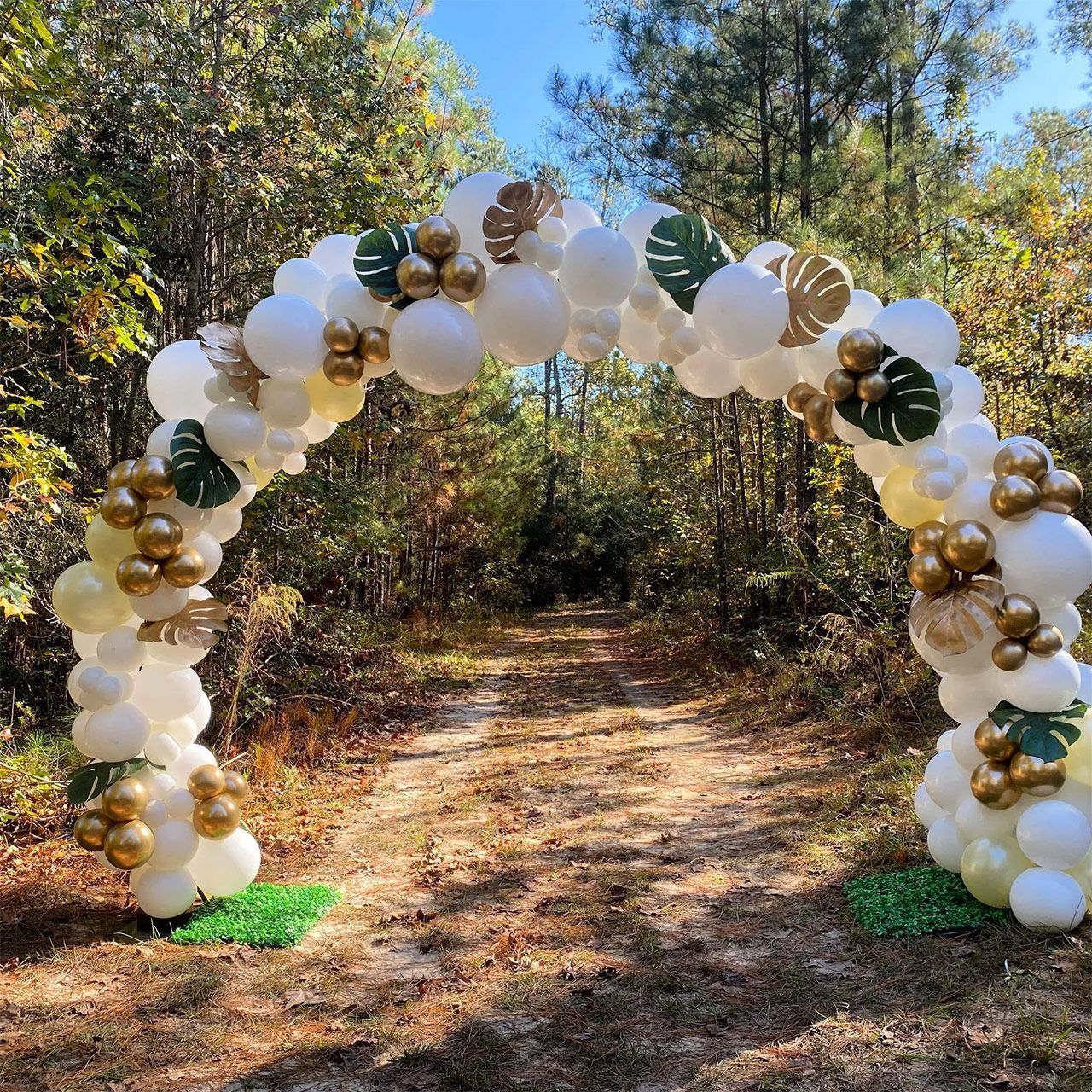 White, gold, and green balloon archway over a path in a wooded area.