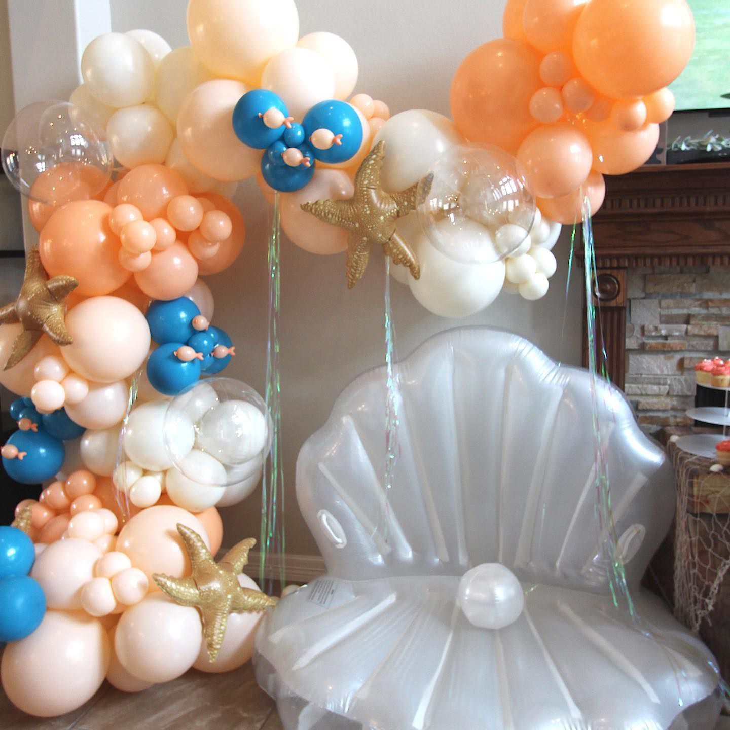 Mermaid-themed party setup: shell chair with balloon arch in coral, peach, white, and blue. Gold starfish accents.