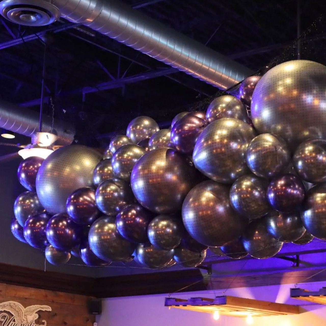 Cluster of silver and purple metallic balloons suspended from a ceiling, lit with purple light.