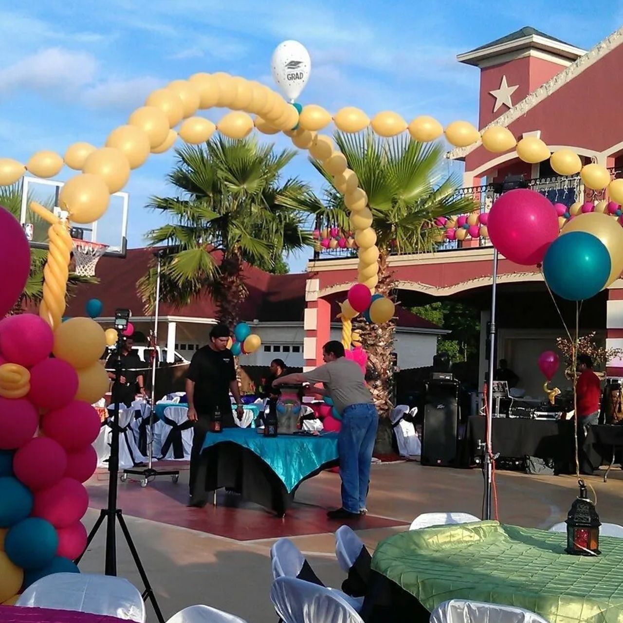 Outdoor event with balloon arch, tables, people, and building with palm trees.