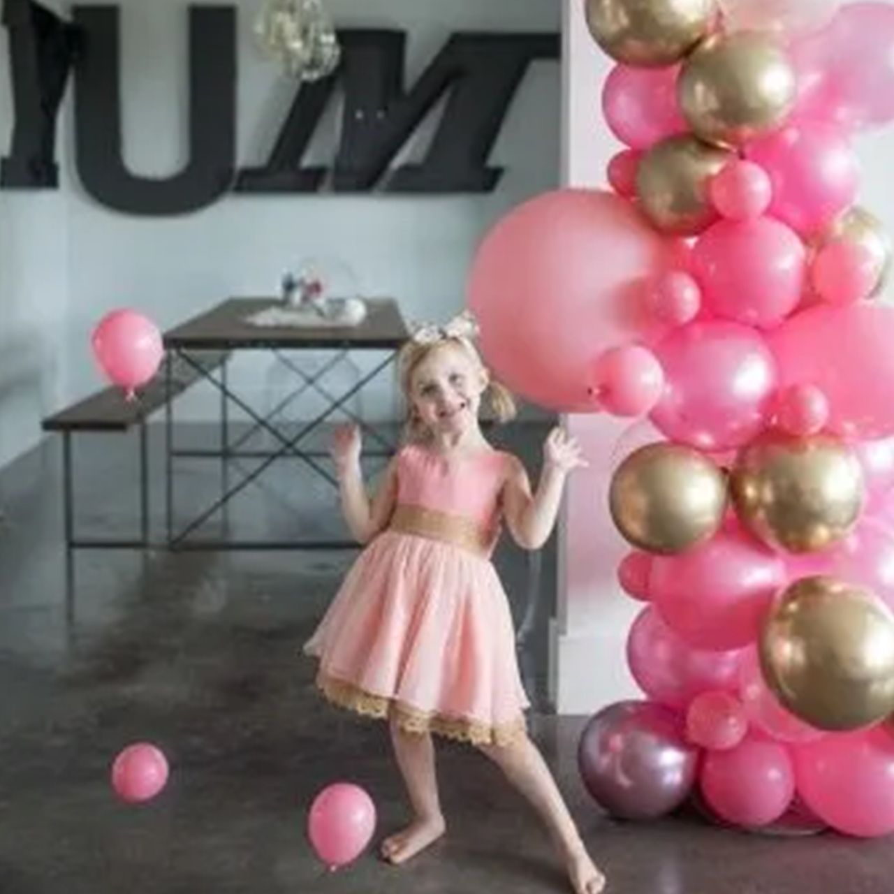Girl in pink dress smiles near a balloon arch with gold and pink balloons.