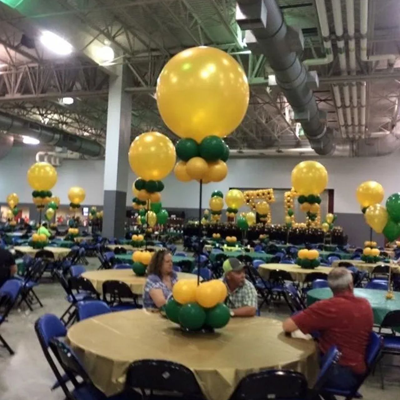 Event hall with round tables decorated with gold and green balloons. People seated at tables.