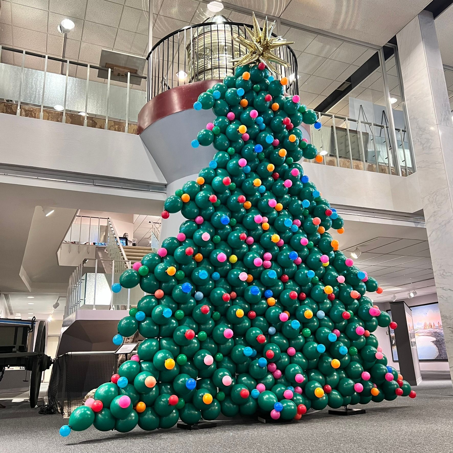 Christmas tree sculpture made of green balloons, decorated with colorful ornaments, inside a building.