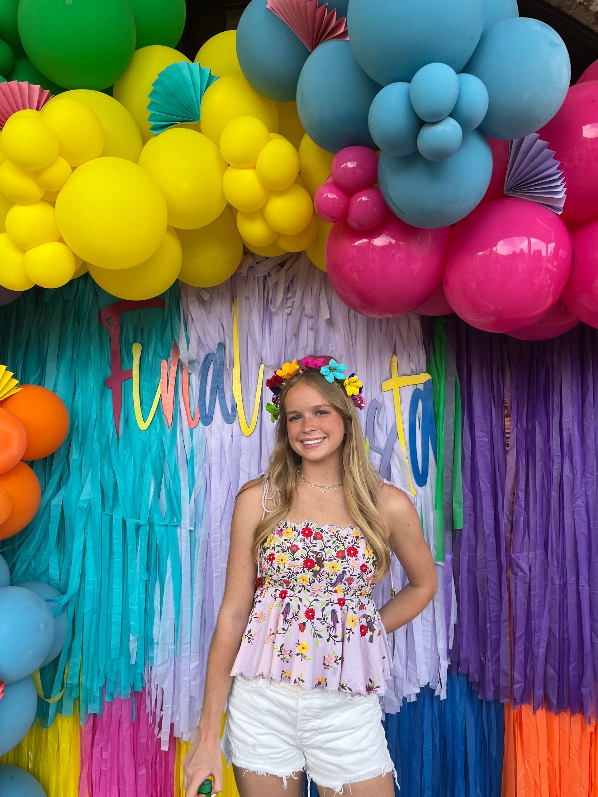 Young person smiles, wearing flower crown and floral top, in front of a colorful backdrop with balloons.
