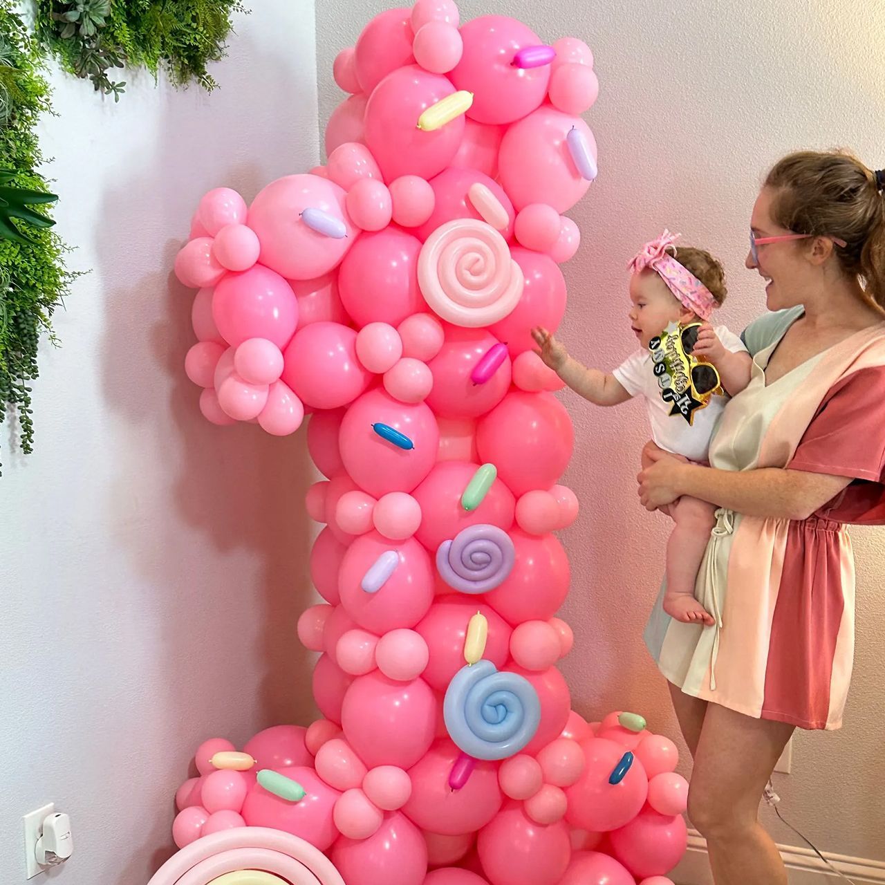 Woman holding baby near pink balloon number one. Party decorations include candy accents.