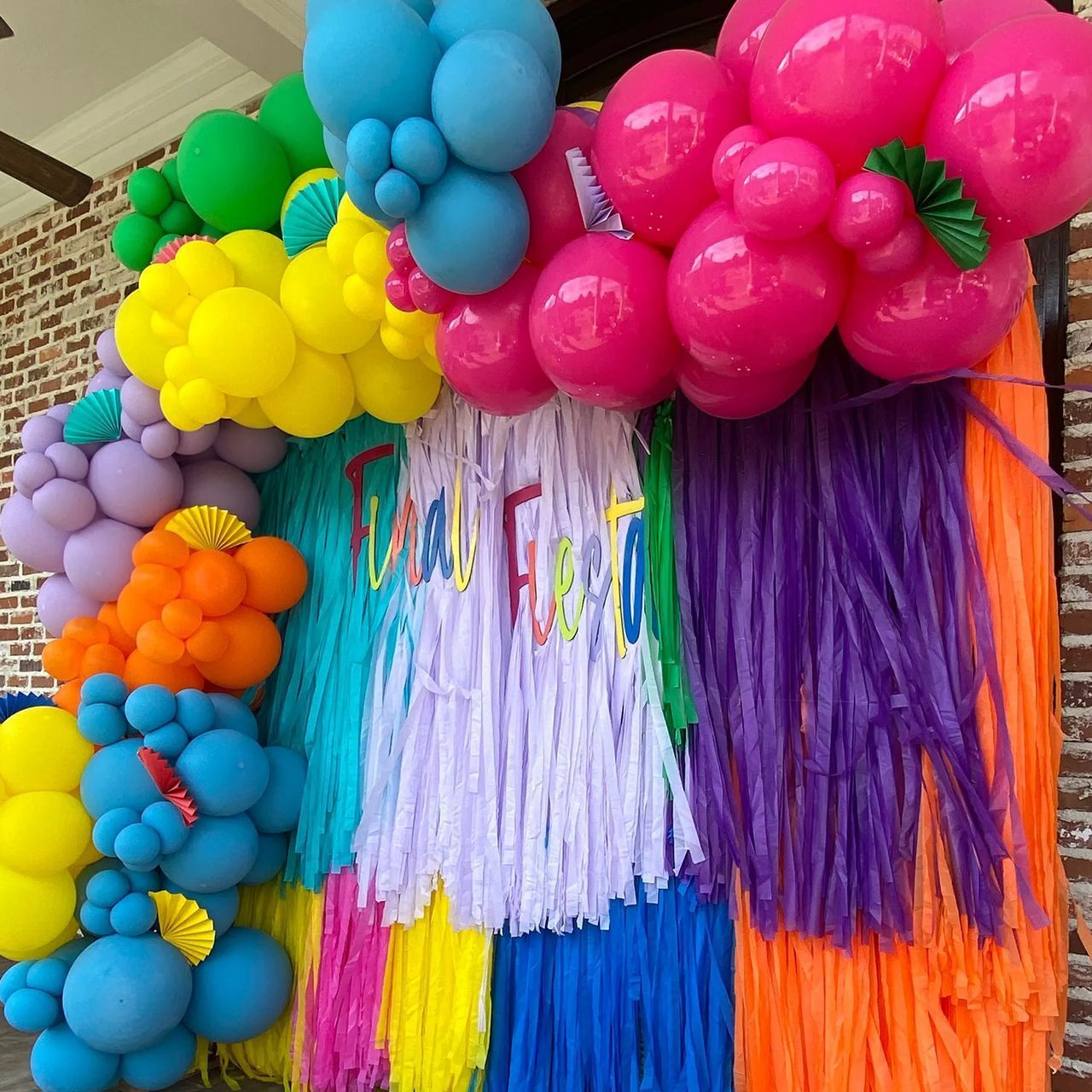 Colorful balloon arch with fringe backdrop, text reads 