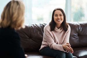 Two people sit on a couch in a bright room, one smiling and talking with the other.