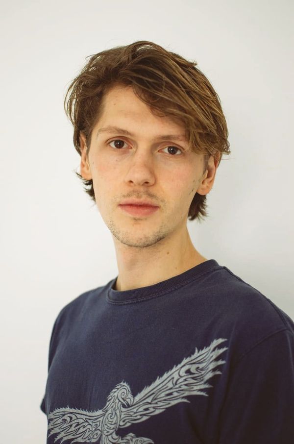 A young man wearing a blue shirt with an eagle on it is standing in front of a white wall.