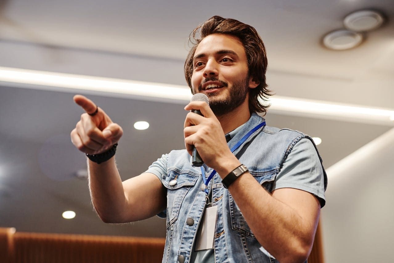 A man with a beard is holding a microphone and pointing.