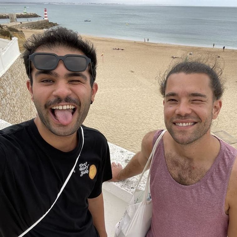 Two men are standing next to each other on a beach and one has his tongue out