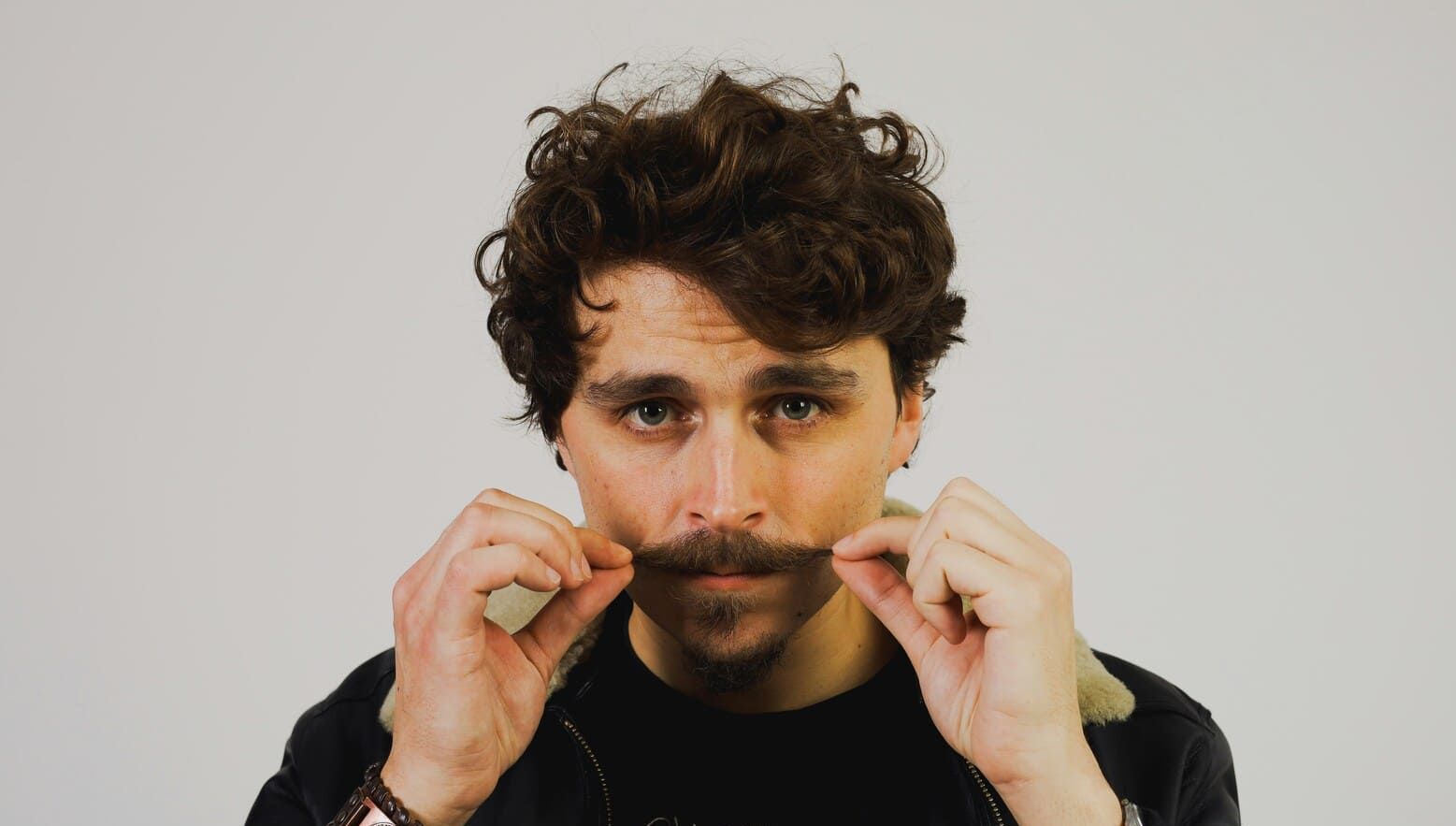 A man with curly hair is holding his mustache in front of his face.