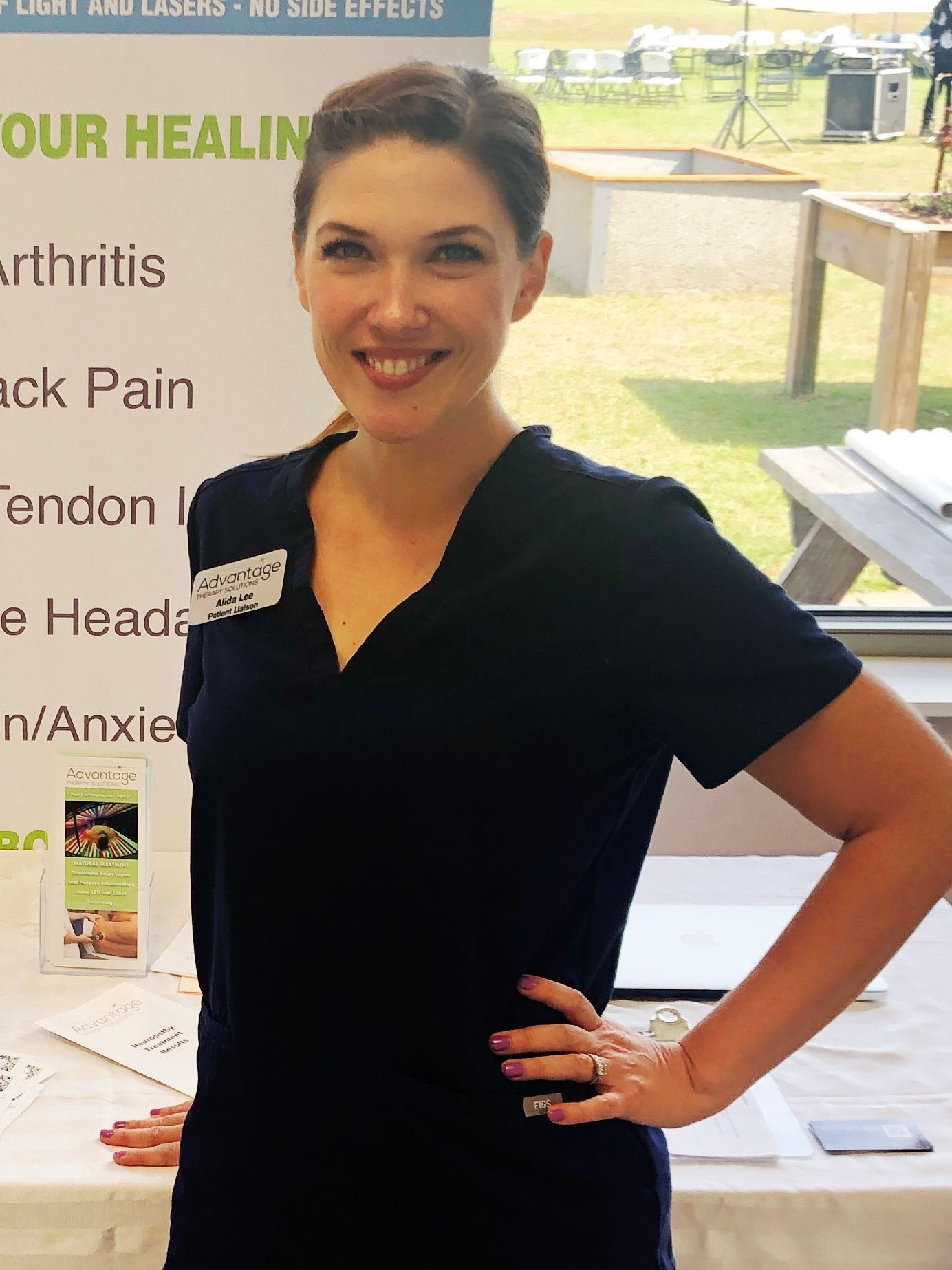 A smiling person in dark blue scrubs stands in front of a health service informational banner at an outdoor event.