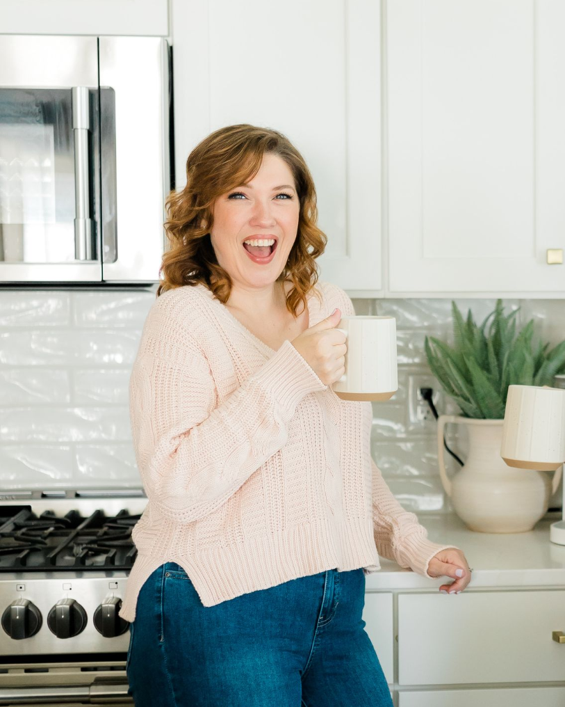 A smiling person in a light pink sweater holds a mug in a white kitchen.