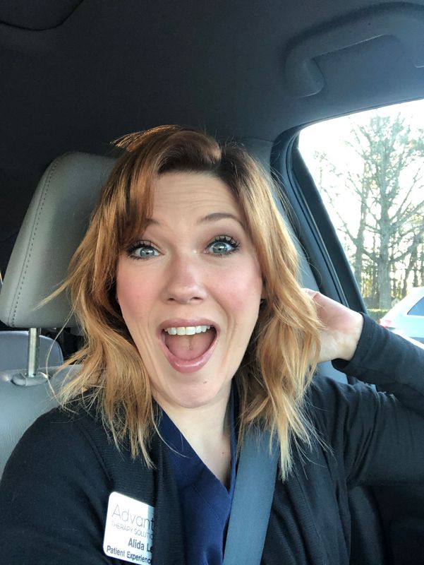 A person with shoulder-length reddish-brown hair, wearing a dark cardigan, smiling with an open mouth inside a car.