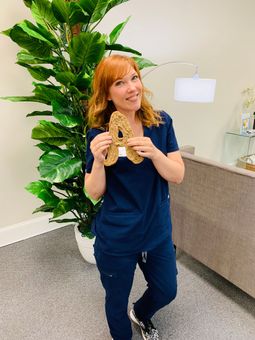 A person wearing dark blue scrubs stands in an office, smiling while holding a textured letter A.