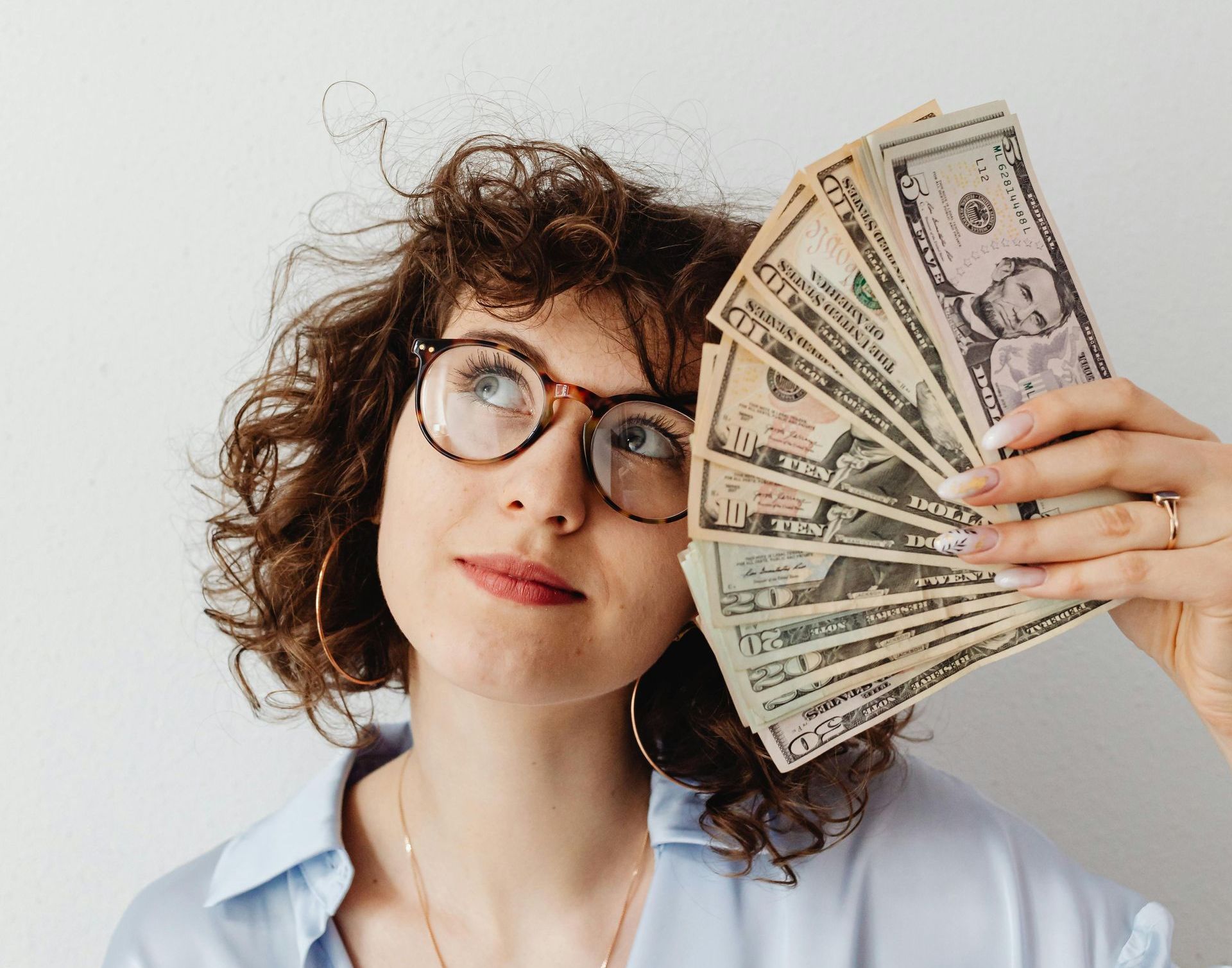 A woman wearing glasses is holding a fan of money in front of her face.
