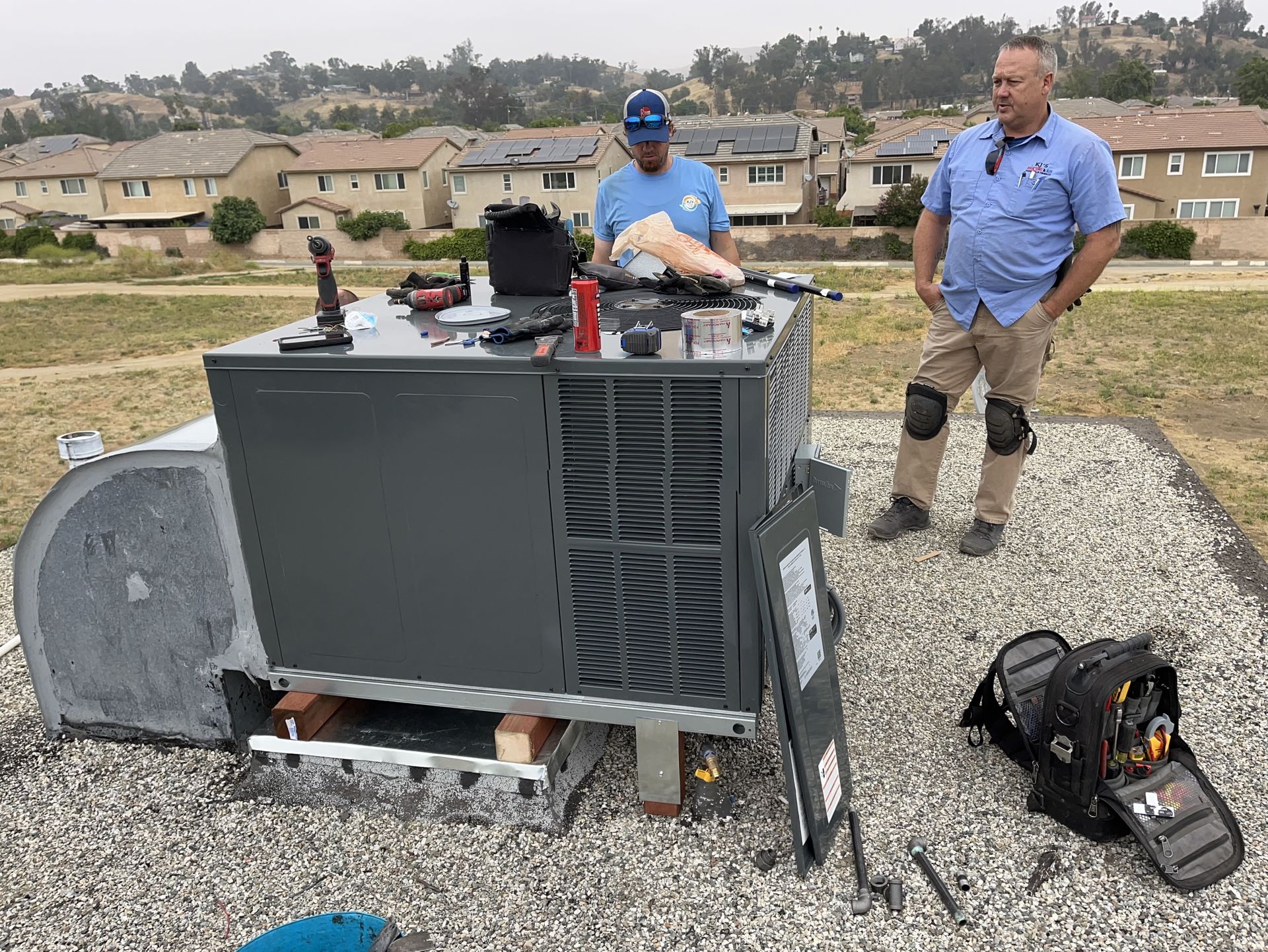 Two men are working on an air conditioner on the roof of a building.
